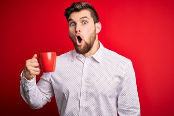 Young blond businessman with beard and blue eyes drinking red cup of coffee scared in shock with a surprise face, afraid and excited with fear expression