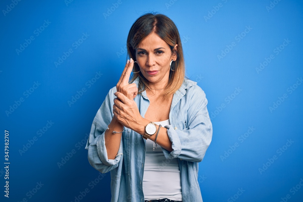 Middle age beautiful woman wearing casual shirt standing over isolated blue background Holding symbolic gun with hand gesture, playing killing shooting weapons, angry face