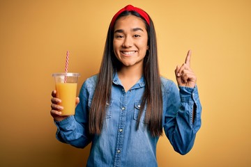 Poster - Young beautiful asian woman drinking healthy glass of orange juice over yellow background surprised with an idea or question pointing finger with happy face, number one