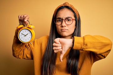 Canvas Print - Young asian woman holding vintage alarm clock standing over isolated yellow background with angry face, negative sign showing dislike with thumbs down, rejection concept