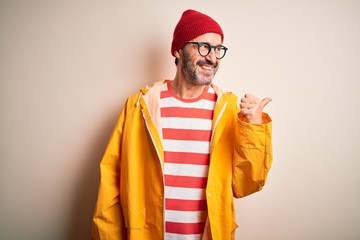 Wall Mural - Middle age hoary man wearing glasses and rain coat standing over isolated white background smiling with happy face looking and pointing to the side with thumb up.