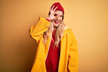 Poster - Young beautiful blonde woman wearing raincoat and wool cap over isolated yellow background doing ok gesture with hand smiling, eye looking through fingers with happy face.