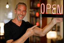Open For Business Concept, Small Business Owner Reopening Premises After Covid-19 Virus Pandemic, Happy Man Pointing To Red Neon Open Sign At A Bar Restaurant Or Cafe Window After Coronovirus Lockdown