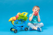 © evakerrigan - A hungry blond child sits near a supermarket basket full of food carts and bites a huge red apple. The concept of food delivery and healthy eating of fruits and vegetables.