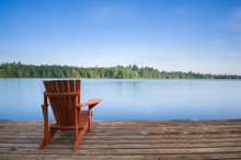 Adirondack Chair Sitting On A Wood Dock Facing A Calm Lake. Across The Water There Are Green Trees.