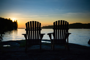 Silhouette of two Adirondack chairs sit on a rock formation facing the calm waters of a lake during a sunset in Muskoka, Ontario Canada. 