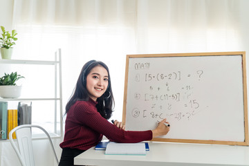 Asian school young woman teacher working from home teach online math subject to student studying from home. Girl writes on whiteboard, talk on headphone. Remote education class during covid19 pandemic