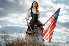 Girl In Historical Dress Of 18th Century With Flag Of United States. July 4 Is US Independence Day. Woman Of Patriot Freedom Fighter In Outdoor On Background Cloudy Sky
