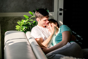 Lovers relax on a cozy sofa in the living room, a woman lies on the man s shoulder, a man and a woman smile and spend a lazy weekend. The room has bright sunlight and hard shadows.