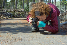 Behind The Scenes Photograph Of A Young, Red-headed Female Photographer Going Low In An Awkward Position To Shoot A Small Garter Snake On A Paved Path In A Wooded Area On A Cold Sunny Autumn Day.