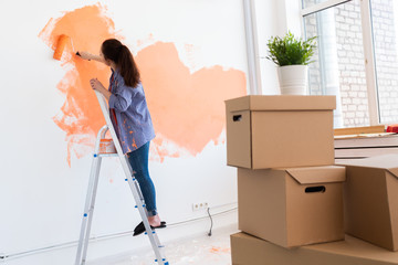 Happy young woman painting interior wall with paint roller in new house. A woman with roller applying paint on a wall.