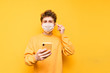 © bodnarphoto - A young man in a protective mask stands on a yellow background with a smartphone in his hand, listens to music in wireless headphones and looks at the camera. Coronavirus pandemic. COVID-19