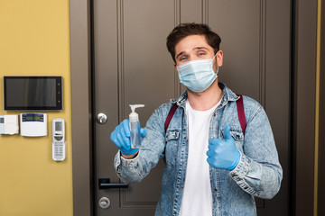 Man in medical mask and latex gloves showing thumb up and holding hand sanitizer near boor at home