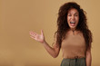 Angry young curly dark skinned brunette woman with casual hairstyle raising emotionally her hand while screaming madly, isolated over beige background