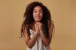 Portrait of cute young glad long haired curly dark skinned female with natural makeup folding raised hands and looking positively at camera, isolated over beige background