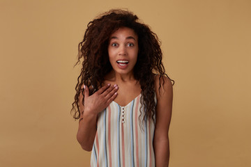 Excited young lovely brown haired dark skinned brunette woman with voluminous hairstyle raising surprisedly eyebrows and keeping palm on her chest while posing over beige background