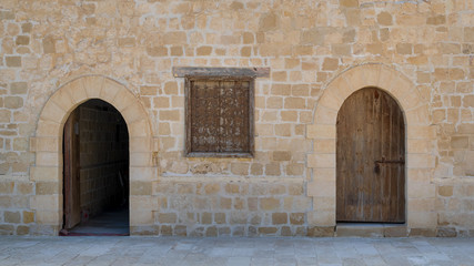 Two weathered wooden arched doors and one closed rusted wrought iron window on bricks stone wall and tiled stone floor