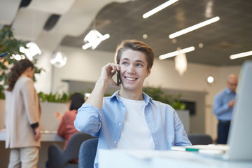 Wall Mural - portrait of smiling young man lspeaking by smartphone while working with laptop in office, copy spac