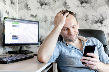 young male freelancer at home at a computer with a smartphone in his hand