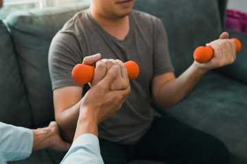 Asian physiotherapist helping a patient lifting dumbbells work through his recovery with weights in clinic room.