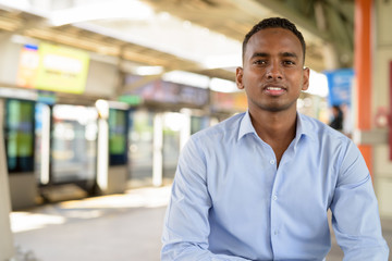 Wall Mural - Happy young handsome African businessman smiling while sitting at the sky train station