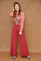 Full-length vertical portrait of a young Caucasian red-haired woman on a beige background in various poses. Nice girl model dressed in red pants and a pink blouse.