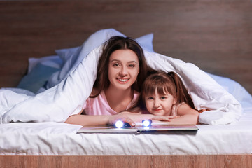 woman and her little daughter reading book under blanket at night