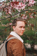 © KseniaJoyg - A young man with blond hair in a brown jacket and a backpack walks in a blooming garden, spring blooming pink cherry trees, close-up portrait