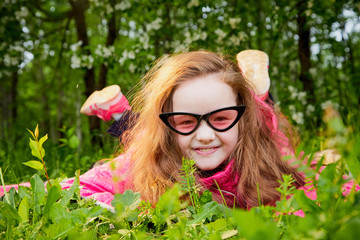 Playful funny little girl with long hair and in pink glasses having rest and fun on green grass in a summer park