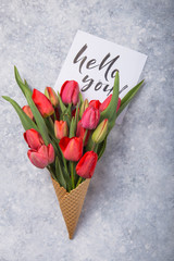 Wall Mural - red  beautiful tulips in an ice cream waffle cone with card Hello you on a concrete background. Conceptual idea of a flower gift. Spring mood