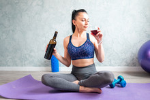 Young Woman In Sport Outfit  Doing Fitness Exercise With Dumbbell And Drinking Glass Of Red Wine, Sitting On Fitness Ball At Home