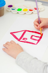 Wall Mural - A hand of girl with brush drawing house of dream by watercolor on white sheet of paper. Kid draws house.