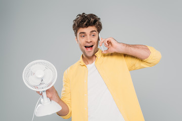 Front view of excited man holding desk fan and talking on smartphone isolated on grey