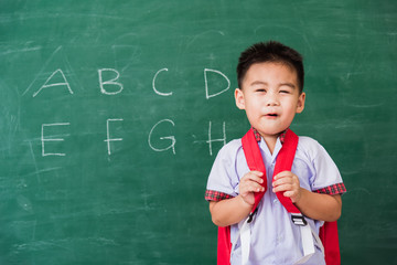 Back to School. Happy Asian funny cute little child boy kindergarten preschool in student uniform wearing school bag stand smiling on green school blackboard, First time to school education concept