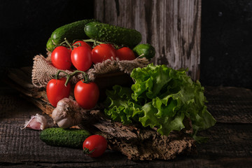 Fresh ripe red tomatoes on a branch, cucumbers, lettuce, garlic on an old wooden board. Ingredients for salad. Dark food photo.