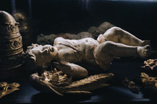 An Ancient Antique Broken Figure Of A Cupid With Broken Gilded Wings Lies On A Black Background Near The Tiara Of The Pope. The Cultural Heritage Of Classical Art Made From White Marble.