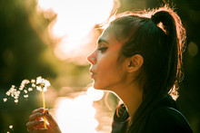 Young Beautiful Woman Blowing Dandelion At Sunset