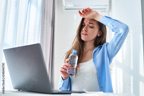 Cuadros en Lienzo Sweating working woman suffering from heat, hot weather and thirst cools down with refreshing cold water bottle during online remote work at computer at home