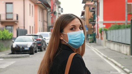 Wall Mural - COVID-19 Young woman with surgical mask walks in city street turns and looks towards the camera