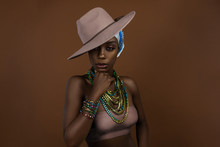 A Fierce Looking Young Black Female With Beautiful Makeup Posing By Herself In A Studio Wearing A Beige Strapless Top, Head Scarf, Beige Hat And Gorgeous Colorful Jewelry.