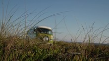 Panoramic Shot Of Grass In The Foreground With A Green Vintage Camping Van Passing Behind On The Edge Of A Cliff
