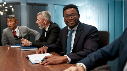 Portrait of handsome african man in formal wear looking at camera and smiling while sitting at the office table with his co workers