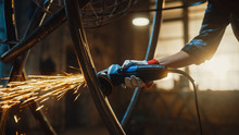 Close Up Of Hands Of A Metal Fabricator Wearing Safety Gloves And Grinding A Steel Tube Sculpture With An Angle Grinder In A Studio. Working With A Handheld Power Tool In A Workshop.