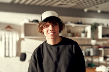 Streetwear. Stylish guy smiling at camera while posing in custom apparel, black baseball cap and sweatshirt. Young man working at custom T-shirt, clothing printing company