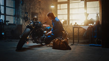 Young Beautiful Female Mechanic Comes To Garage And Starts Working On A Custom Motorcycle. Talented Girl Wearing A Blue Jumpsuit. She Uses A Spanner To Tighten Nut Bolts. Creative Authentic Workshop.