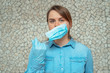 A young blonde woman in a blue shirt wearing blue protective gloves puts a protective mask on her face