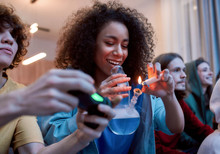 For Great Mood. Young Afro American Girl Lighting Marijuana In The Glass Bong, Relaxing With Friends On The Sofa At Home. Young People Playing Video Games And Smoking Weed