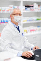 Wall Mural - medicine, healthcare and people concept - senior apothecary wearing face protective medical mask for protection from virus disease at pharmacy cash register