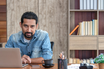 Young Asian businessman in casual clothes stitting and replying an e-mail. Handsome 30s man working at home, looking to camera.