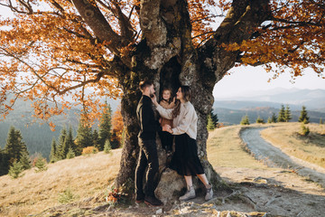 Wall Mural - Stylish young family in the autumn mountains. A guy and a girl with their daughter stand under a large old tree against the background of a forest and mountain peaks at sunset